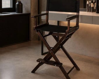 May include: A tall director's chair featuring a dark brown wooden frame and black fabric seat and backrest. The chair has a folding X-frame base, two armrests, and a horizontal footrest bar positioned between the front legs for support.
