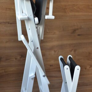 May include: White wooden folding chair with a black padded seat and backrest. The chair is designed with a cross-legged frame and is shown in two sizes, one folded and one partially unfolded.