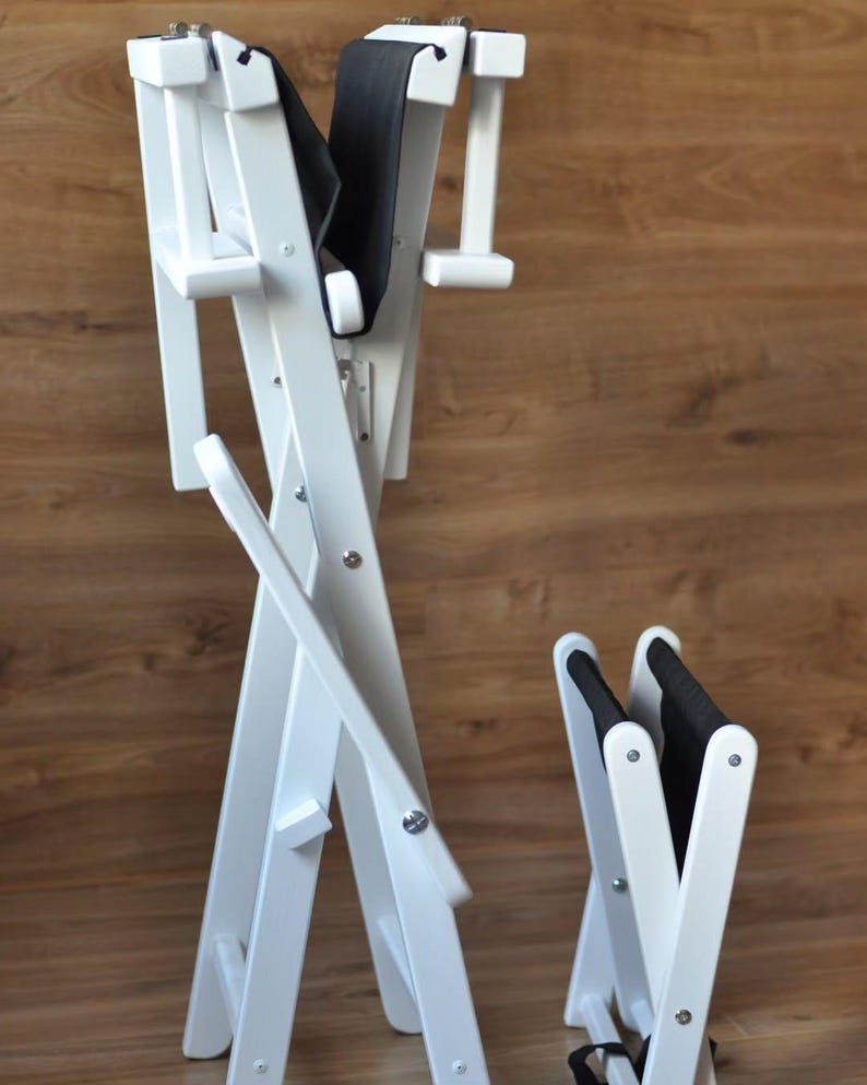May include: White wooden folding chair with a black padded seat and backrest. The chair is designed with a cross-legged frame and is shown in two sizes, one folded and one partially unfolded.
