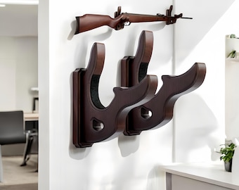May include: Two dark brown wooden gun racks mounted on a white wall. The racks have a decorative, curved design and are designed to hold rifles or shotguns.