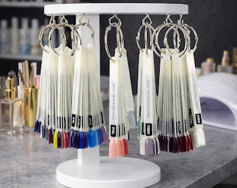 May include: A white nail polish display stand with multiple color swatch fans. Each fan is attached to a silver ring and displays a variety of nail polish colors, including pinks, blues, and purples. The text "KODI PROFESSIONAL" is visible on each fan.