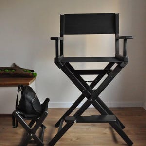 May include: A black director's chair with a matching small folding chair. The director's chair has a high back, armrests, and a footrest. Both chairs are made of wood and have a matte finish. The chairs are on a light brown wooden floor.