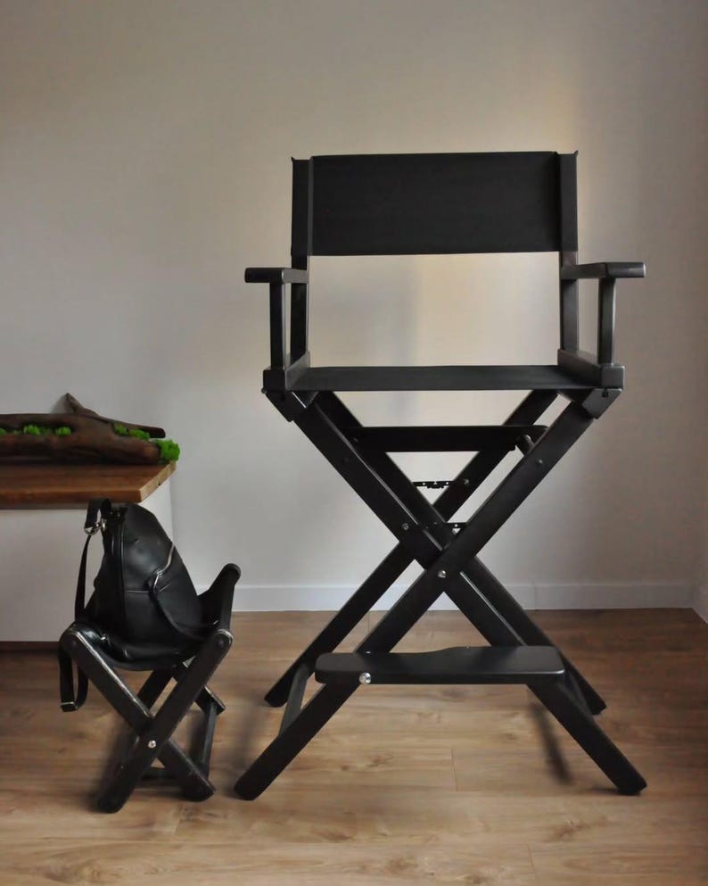 May include: A black director's chair with a matching small folding chair. The director's chair has a high back, armrests, and a footrest. Both chairs are made of wood and have a matte finish. The chairs are on a light brown wooden floor.