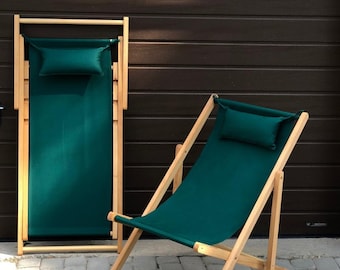 May include: Two wooden deck chairs with teal fabric seats and headrests. One chair is folded, and the other is open, angled slightly. The chairs have a natural wood frame and are set against a dark brown background.