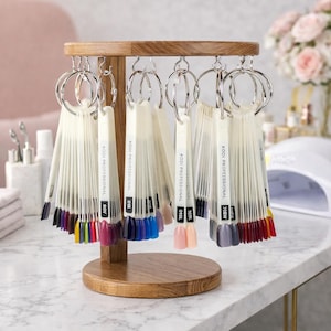 Puede incluir: Un expositor de color de esmalte de uñas de madera con múltiples muestras de color. Cada muestra está etiquetada con un código y unida a un anillo de metal. El expositor es de madera marrón claro y se encuentra sobre una superficie blanca. Los colores de esmalte de uñas incluyen tonos de rosa, azul, morado y rojo.