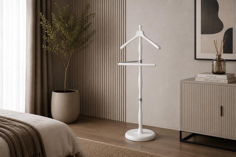 May include: A white wooden valet stand featuring a contoured coat hanger at the top, a horizontal bar for trousers, and a central vertical post supported by a circular base. The design includes silver-toned metal accents connecting the components.