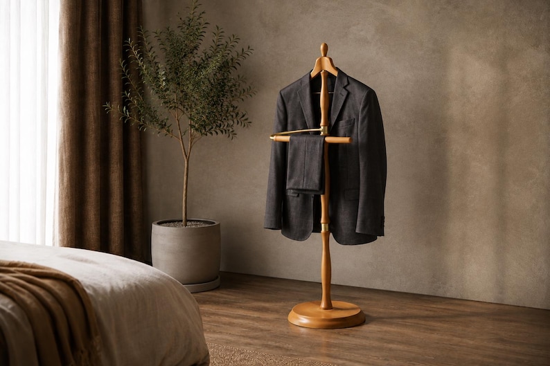 May include: A wooden valet stand with a central pole, a rounded base, and a top hanger section. A dark gray blazer is draped over the hanger, and a pair of matching dark gray trousers is folded over the horizontal bar located below the jacket.