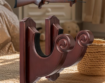 May include: A dark brown wooden gun rack with a decorative scroll design. The rack is designed to hold a rifle, with a matching wooden rifle mounted above. The rack is designed to be mounted on a wall.