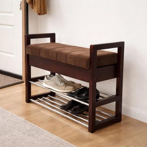 Puede incluir: Un banco zapatero de madera marrón oscuro con un cojín de asiento rectangular de tela marrón acolchada. La base incluye dos niveles horizontales de varillas metálicas plateadas para guardar zapatos, sobre un marco resistente.
