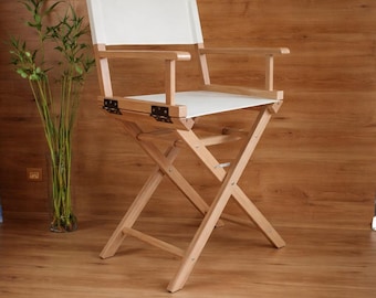 May include: A wooden director's chair with a white canvas seat and back. The chair has a natural wood finish and black metal hinges. The chair is set against a wood-paneled wall and floor.