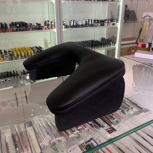 Könnte beinhalten: Schwarze, U-förmige Armlehne für Nageldesigner. Die Armlehne besteht aus einem glatten, dunklen Material und dient zur Unterstützung des Arms des Kunden während der Nagelbehandlungen. Sie steht auf einer Glasoberfläche.