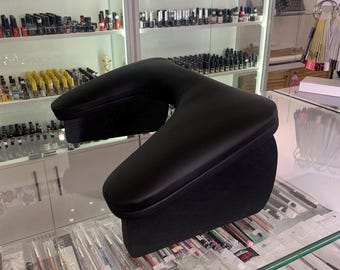 May include: Black, U-shaped armrest for nail technicians. The armrest is made of a smooth, dark material and is designed to support the client's arm during nail services. It is placed on a glass surface.