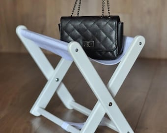 May include: A black quilted leather handbag with a silver chain strap rests on a small, white folding stool. The stool has an X-frame design with a white fabric seat and is made of white painted wood.