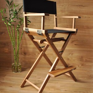 May include: A wooden director's chair with a black seat and backrest. The chair has a folding design with a footrest and armrests. The frame is made of light-colored wood, and the chair is set against a wooden background.