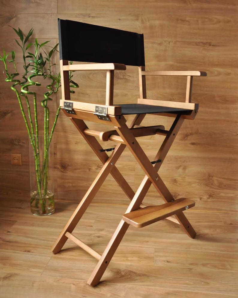 May include: A wooden director's chair with a black seat and backrest. The chair has a folding design with a footrest and armrests. The frame is made of light-colored wood, and the chair is set against a wooden background.