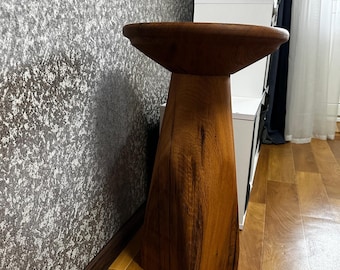 May include: A handcrafted wooden pedestal table with a wide, shallow bowl-shaped top. The pedestal has a geometric, trapezoidal base and a natural wood grain finish. The table is designed for decorative use.