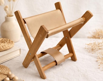 May include: A small, folding wooden magazine rack with a light tan leather seat and handle. The wooden frame is a light brown color. The rack is designed to hold magazines or books. The background is a soft, neutral color.