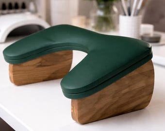 May include: A dark green, U-shaped armrest with a wooden base. The armrest is designed for comfort and support, with a smooth, padded surface. The wooden base provides stability and a natural aesthetic. The armrest is likely used in a beauty or nail salon setting.