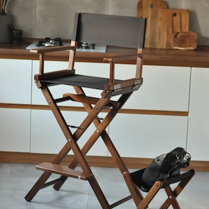 May include: A tall, folding director's chair with a dark brown wooden frame and black canvas seat and back. The chair has a footrest and armrests. A black bag rests on a smaller, matching folding chair.