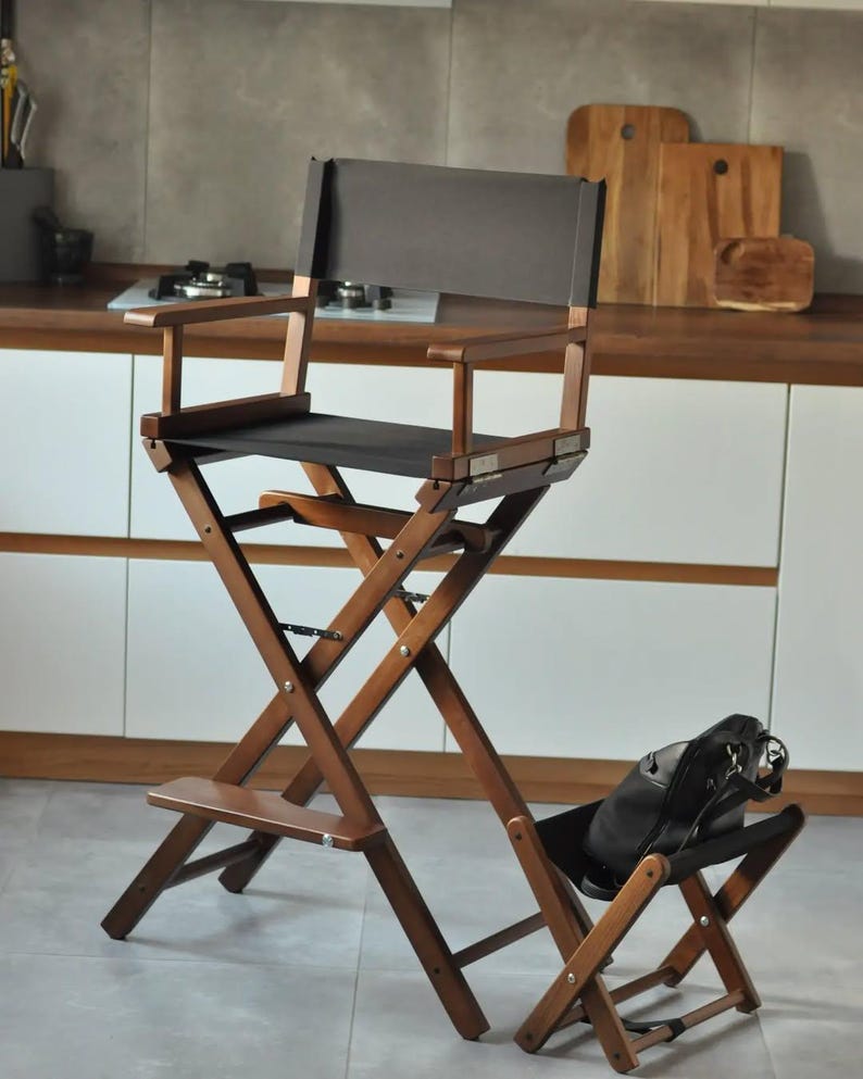 May include: A tall, folding director's chair with a dark brown wooden frame and black canvas seat and back. The chair has a footrest and armrests. A black bag rests on a smaller, matching folding chair.