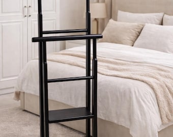 May include: A black wooden valet stand with a hanger, a horizontal bar, and a small shelf. The stand is designed to hold clothing and accessories. It is placed in a bedroom setting with a bed and neutral-toned decor.
