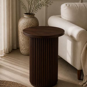 May include: A dark brown, cylindrical side table with a round top. The table's base features vertical, grooved detailing. The table is approximately 20 inches in diameter and 24 inches tall. The table is in a living room setting with a beige sofa and a decorative vase.