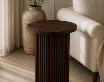 May include: A dark brown, cylindrical side table with a round top. The table's base features vertical, grooved detailing. The table is approximately 20 inches in diameter and 24 inches tall. The table is in a living room setting with a beige sofa and a decorative vase.