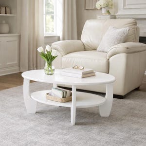 May include: A white oval coffee table with a lower shelf, holding a woven basket with books. The table has a vase of white tulips and a stack of books with glasses on top. A cream-colored leather armchair is in the background.