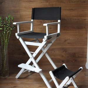 May include: A white director's chair with a black seat and backrest, and a matching footstool. The chair has a folding design and is made of wood. The background is a wooden wall.