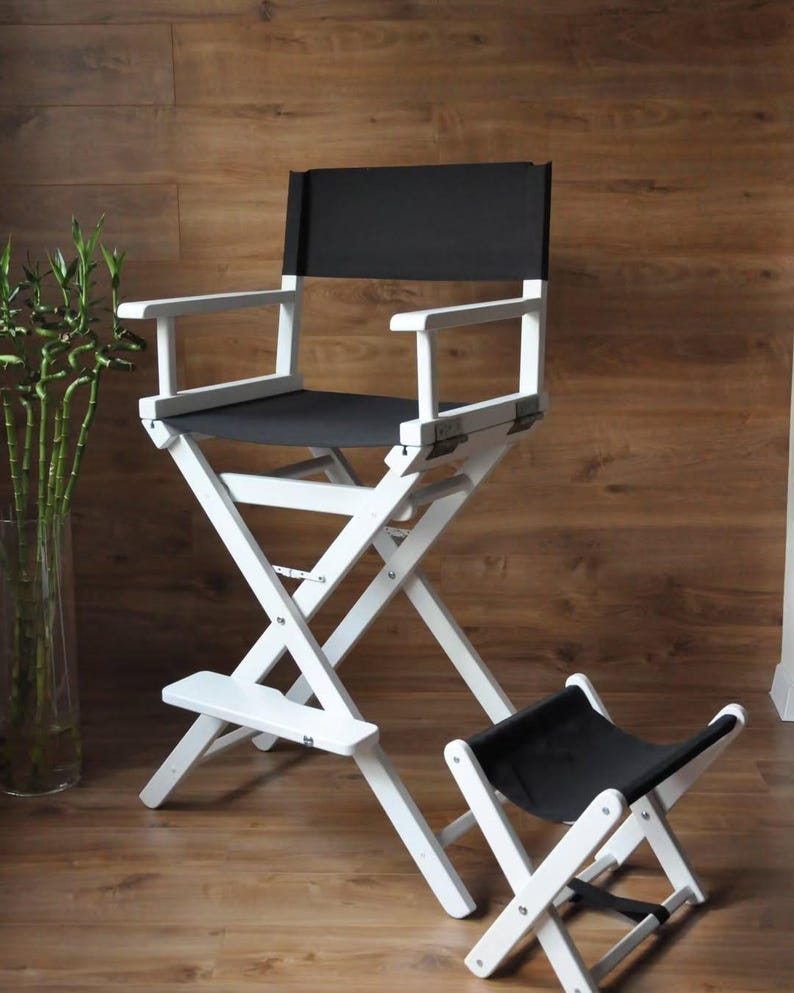 May include: A white director's chair with a black seat and backrest, and a matching footstool. The chair has a folding design and is made of wood. The background is a wooden wall.