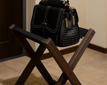 May include: A black leather handbag with gold-colored studs and hardware sits on a dark brown wooden folding luggage rack. The bag has two zippered compartments and a top handle. The rack is made of dark wood and has a cross-legged design.