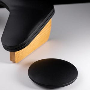 Può includere: Un oggetto nero e arrotondato è posizionato su una superficie bianca, con un supporto in legno e un oggetto nero e curvo sullo sfondo. Il supporto in legno è rettangolare e marrone chiaro. Gli oggetti neri hanno una finitura opaca.