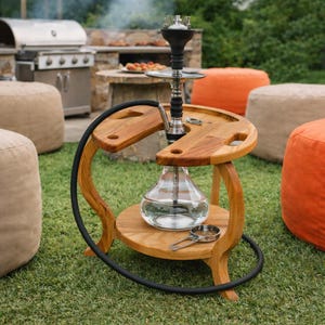 Może przedstawiać: Drewniana fajka wodna z przezroczystą szklaną podstawą i srebrnym metalowym trzonem. Fajka wodna stoi na drewnianym stojaku o zakrzywionym kształcie i czarnym wężu. Stojak ma okrągły blat z wbudowaną tacą.