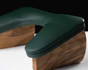 May include: A modern massage table headrest with a dark green, curved cushion and a wooden base. The headrest is designed for comfort and support, with a sleek, minimalist aesthetic. The wooden base has a natural wood grain.