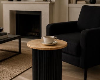 May include: A modern side table with a light wood top and a black, vertically-slatted base. A white teacup and saucer sit on the table. The table is in a living room setting with a black armchair and fireplace.