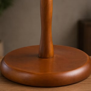 May include: A smooth, medium-brown wooden stand featuring a circular, flat base and a vertical, slightly tapered cylindrical post extending upward from the center. The wood grain is visible across the polished surface of the base and the post.