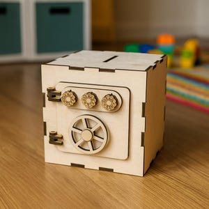 May include: A wooden money box shaped like a safe, with a slot on top for inserting money. The front features a safe door design with a combination dial and a wheel. The box is made of light-colored wood and sits on a wooden floor.