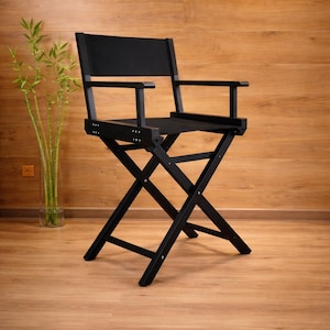 Puede incluir: Silla de director negra con asiento y respaldo de lona, y reposabrazos. La silla tiene un diseño plegable en forma de X y está colocada contra una pared y un suelo de madera. Un jarrón de bambú está a la izquierda.