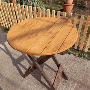 May include: A round, wooden folding table with a light brown tabletop and a dark brown, crisscross base. The table is outdoors, set on a concrete surface, with a small wooden fence and potted plants in the background. The table is approximately 76 cm in diameter.
