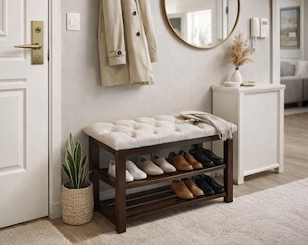 May include: A wooden shoe bench with a tufted, off-white cushion and two shelves filled with shoes. A beige coat hangs above the bench. A potted snake plant sits to the left. A round mirror and a white cabinet are in the background.