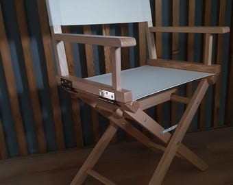 May include: A folding director's chair with a natural wood frame and white canvas seat and back. The chair has armrests and metal hinges. The chair is designed for portability and easy storage.