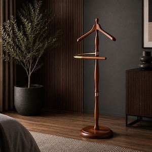 Pode incluir: Um cabideiro de madeira independente com acabamento em castanho escuro, apresentando um cabide contornado no topo e uma barra horizontal para calças. O poste central tem detalhes em metal dourado e assenta numa base circular.