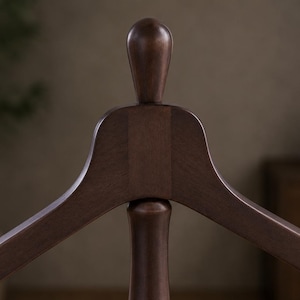 May include: A dark brown wooden clothes hanger featuring a smooth, curved frame and a central vertical post topped with a rounded, finial-like knob. The wood grain is visible across the surface, showcasing a polished, uniform finish throughout.