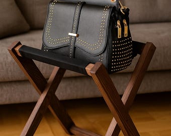 May include: A black leather handbag with gold-colored studs and hardware sits on a wooden folding stool. The stool has a dark wood finish and a black fabric seat. The background features a gray sofa and a wooden floor.