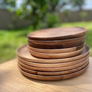 May include: A collection of wooden plates with a natural wood grain. The plates are circular with a raised edge. The wood tones vary from light to dark brown, creating a layered effect. The plates are stacked on a wooden surface.