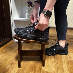 May include: Black athletic shoes with a mesh upper and a chunky sole. The shoes are on a small wooden stool. One shoe is being laced up. The other shoe is on the floor.
