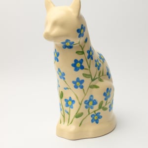 Puede incluir: Una figura de gato de cerámica de color crema con un patrón pintado a mano de flores nomeolvides azules con centros amarillos y tallos verdes. El gato está representado en posición sentada con la cabeza ligeramente girada hacia la izquierda.