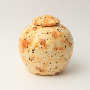 Op de afbeelding: Een ronde keramische pot met deksel, voorzien van een crèmekleurige basis met oranje en gele vlekken en kleine zwarte spikkels. Een subtiele hartvorm is in reliëf aangebracht op de voorzijde van de pot, die een gladde en glanzende afwerking heeft.