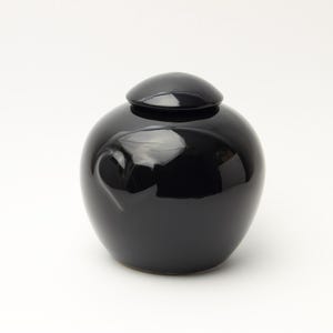 Op de afbeelding: Een glanzende zwarte keramische pot met een rond, bolvormig lichaam en een bijpassend koepelvormig deksel. Het oppervlak van de houder is glad en reflecterend, met een subtiel, ingesprongen hartvormig detail aan de zijkant van het vat.