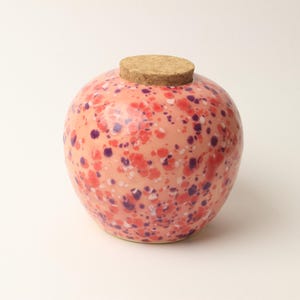Könnte beinhalten: Ein runder Keramiktopf mit Korkdeckel. Der Topf ist pfirsichfarben mit einem gesprenkelten Muster aus roten und violetten Punkten. Der Korkdeckel hat eine hellbraune Farbe. Der Topf steht auf einer weißen Oberfläche.